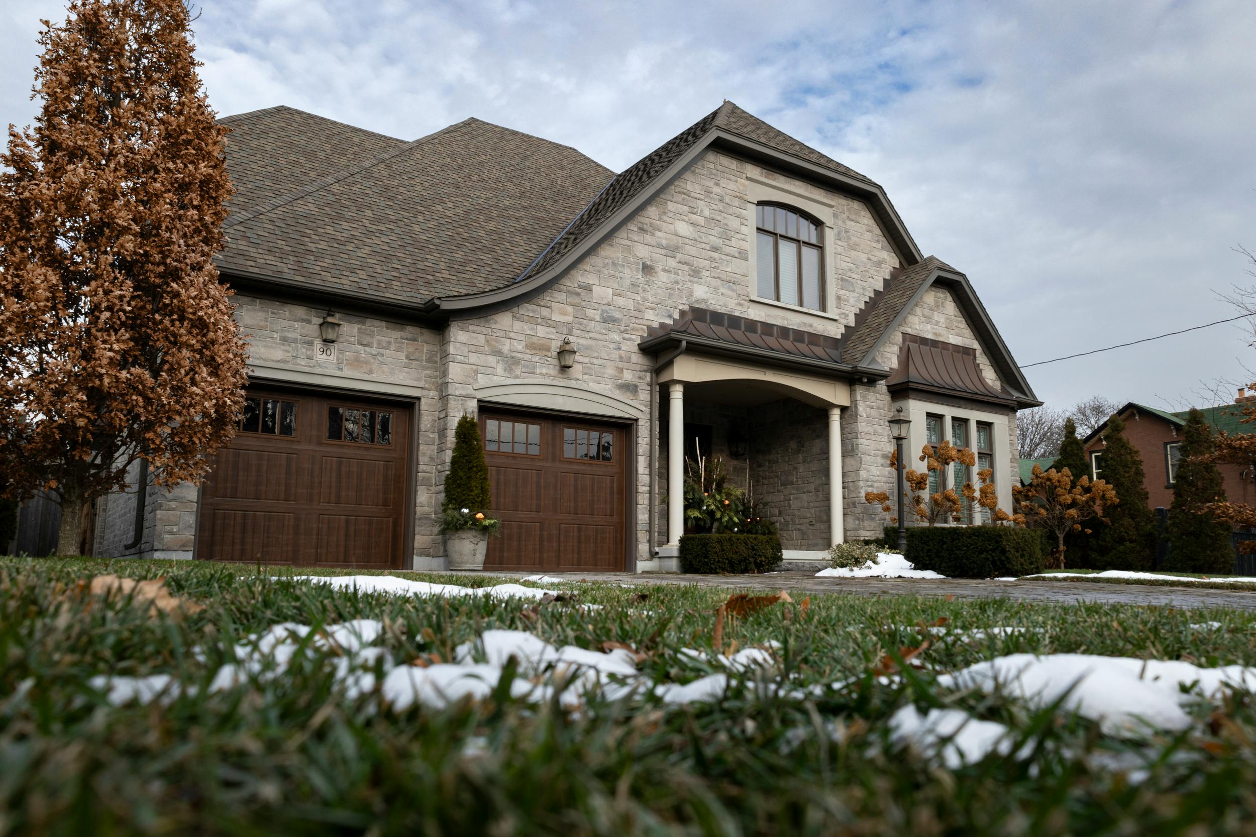 Nos biens Charming stone house with wooden garage doors and a snow-dusted lawn under a cloudy winter sky.