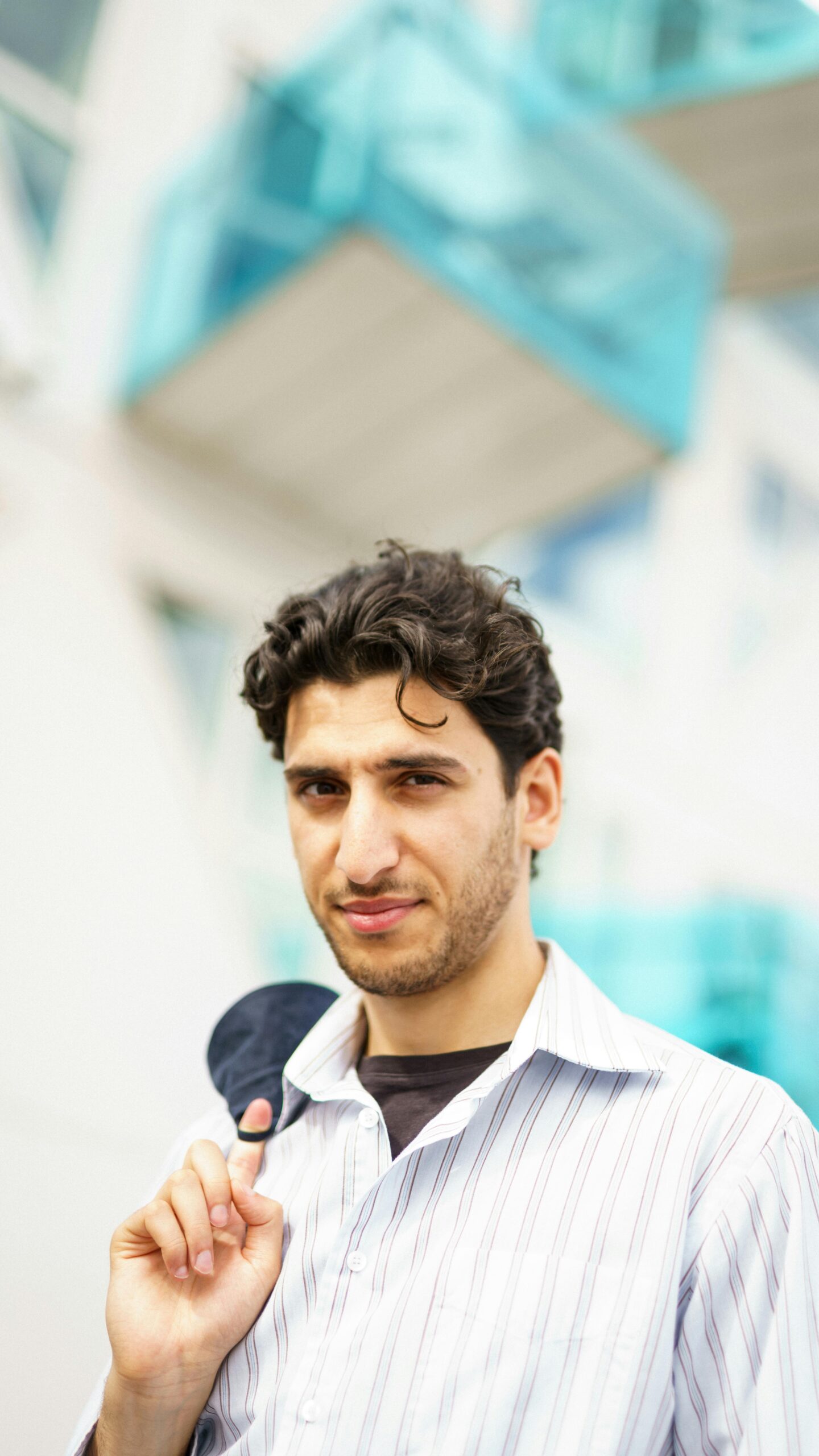 Outdoor portrait of a curly-haired caucasian man holding sunglasses, wearing a striped shirt against a modern building backdrop.