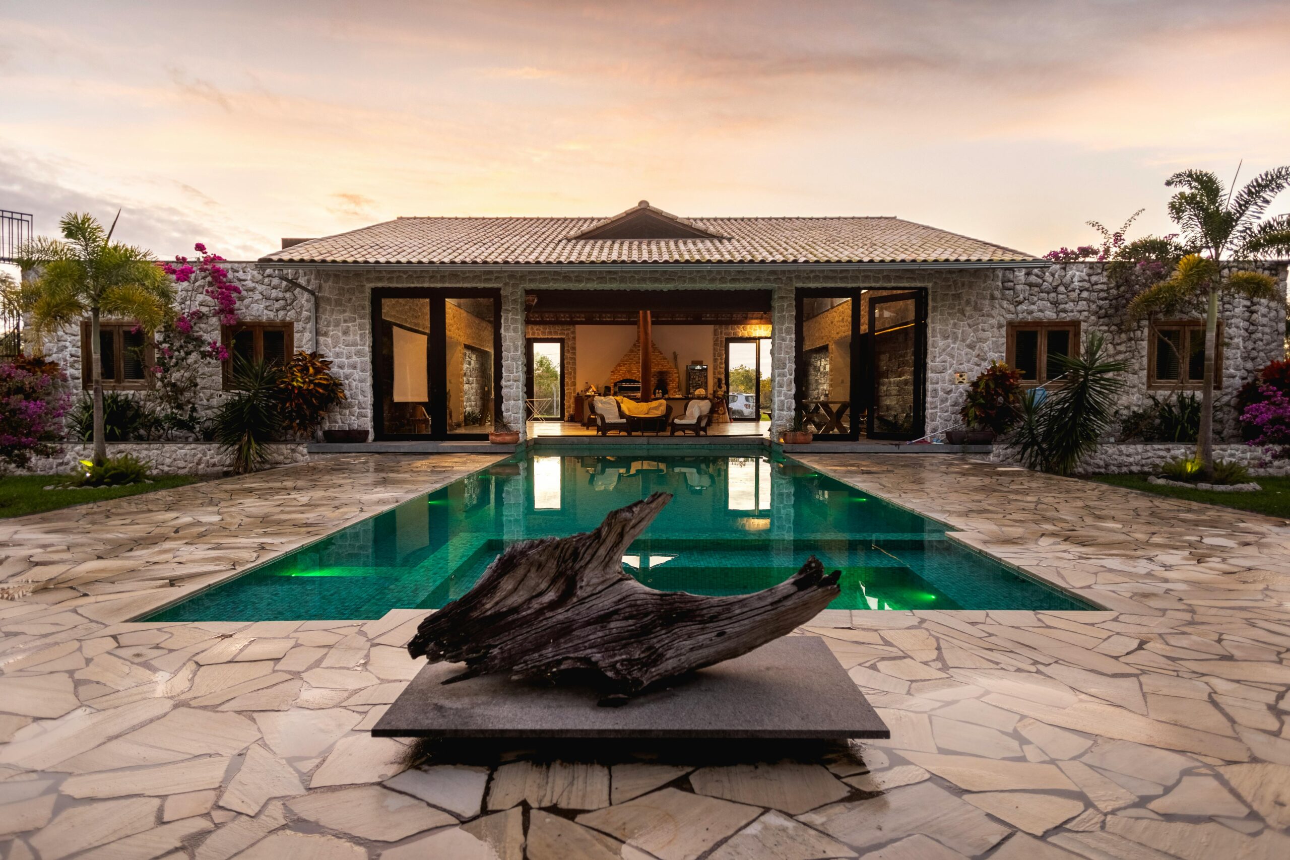Nos biens Elegant villa with a pool, stone facade, and tropical garden at sunset.
