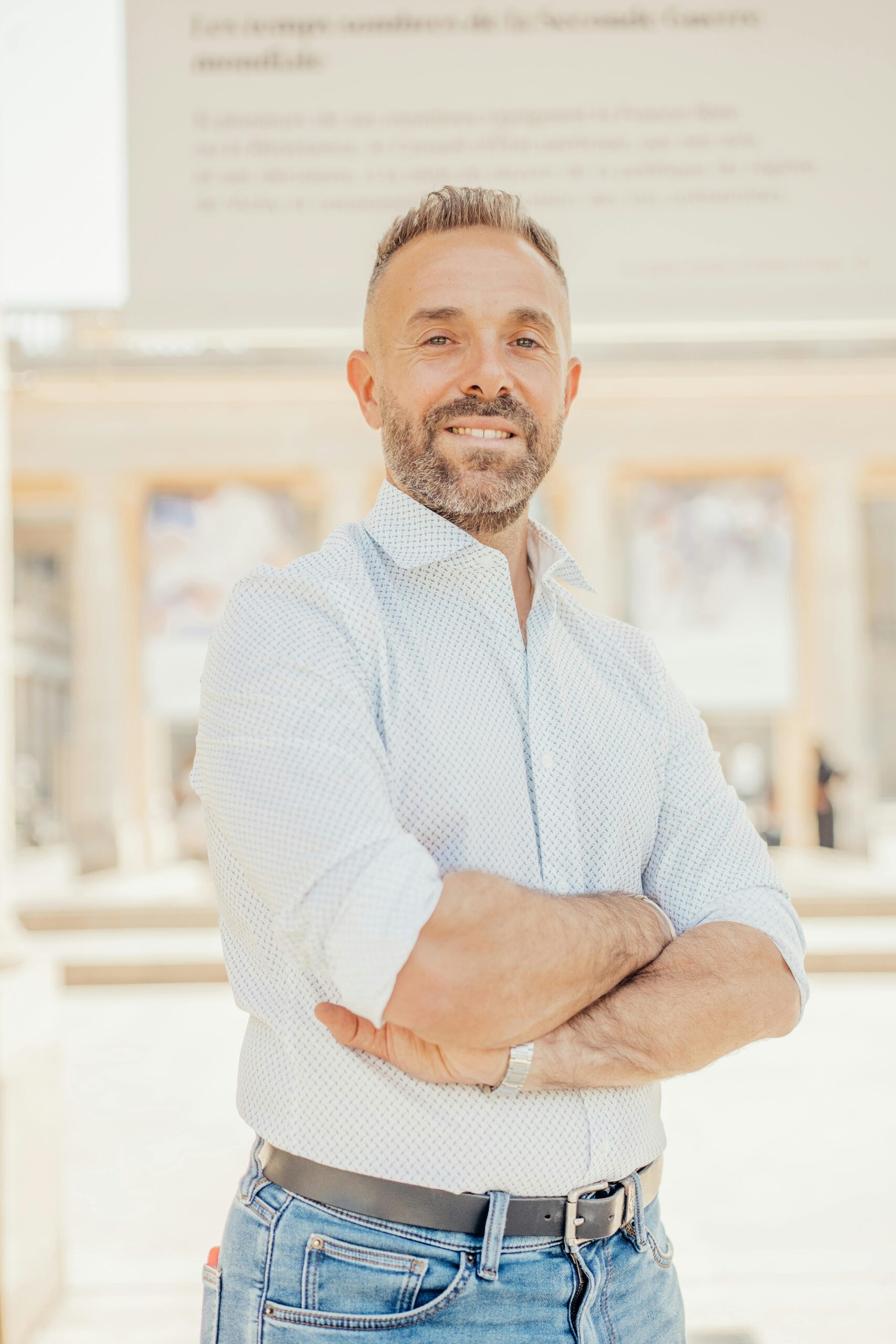 A confident adult man poses in front of Parisian architecture during a sunny day.